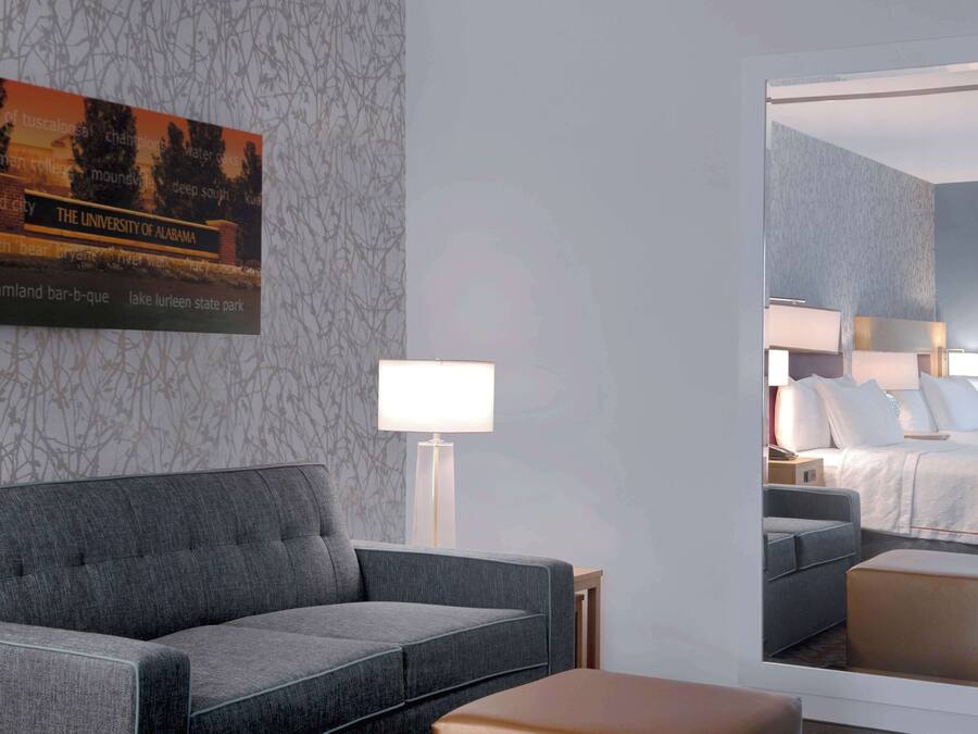 A hotel room with a grey sofa, a bedside table with a lamp, and a large mirror reflecting the room.