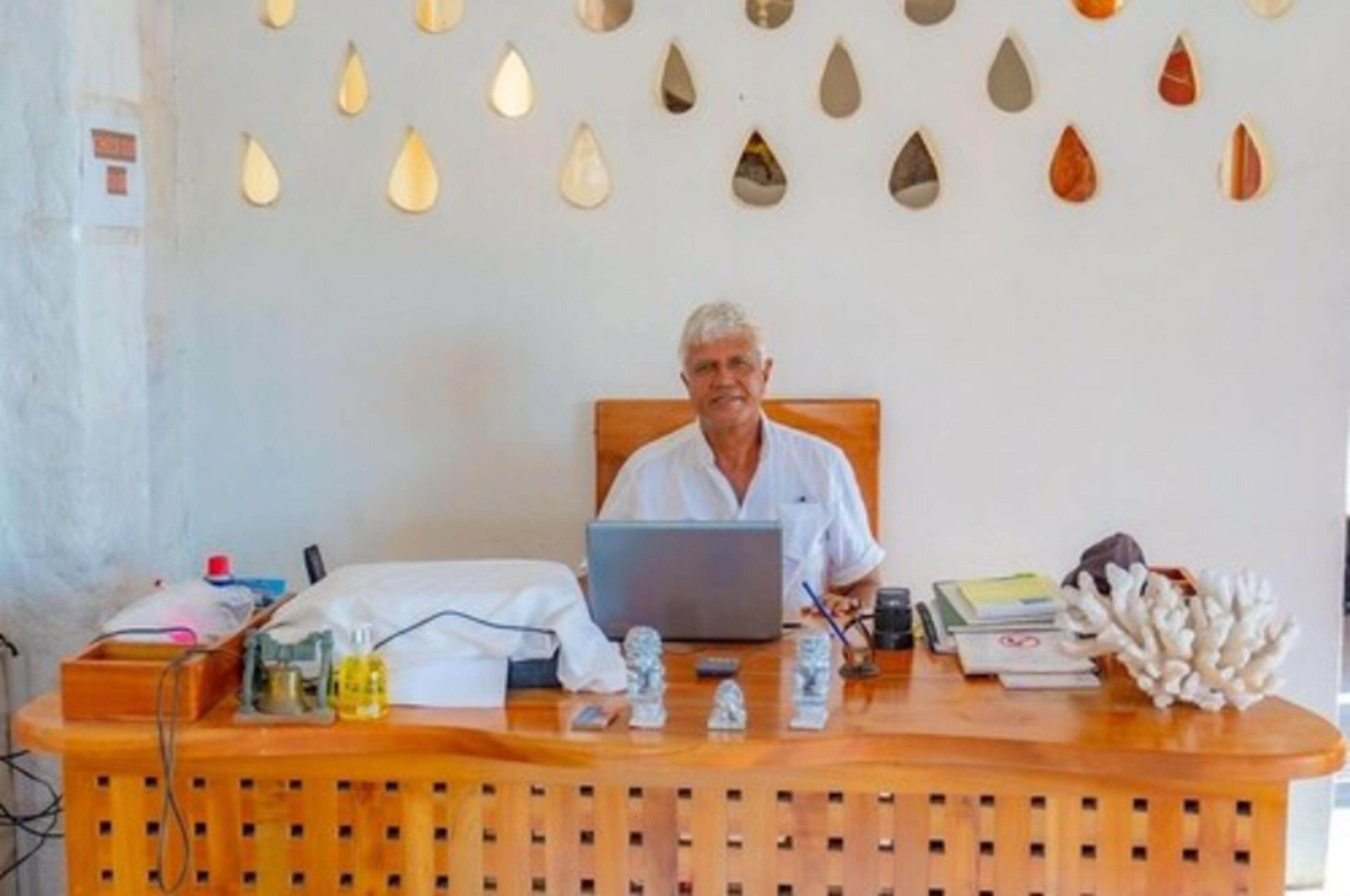 Een receptiebalie met een laptop, een persoon in een wit overhemd zittend erachter, en verschillende items zoals een fles, papieren en een decoratief koraalstuk op de balie.