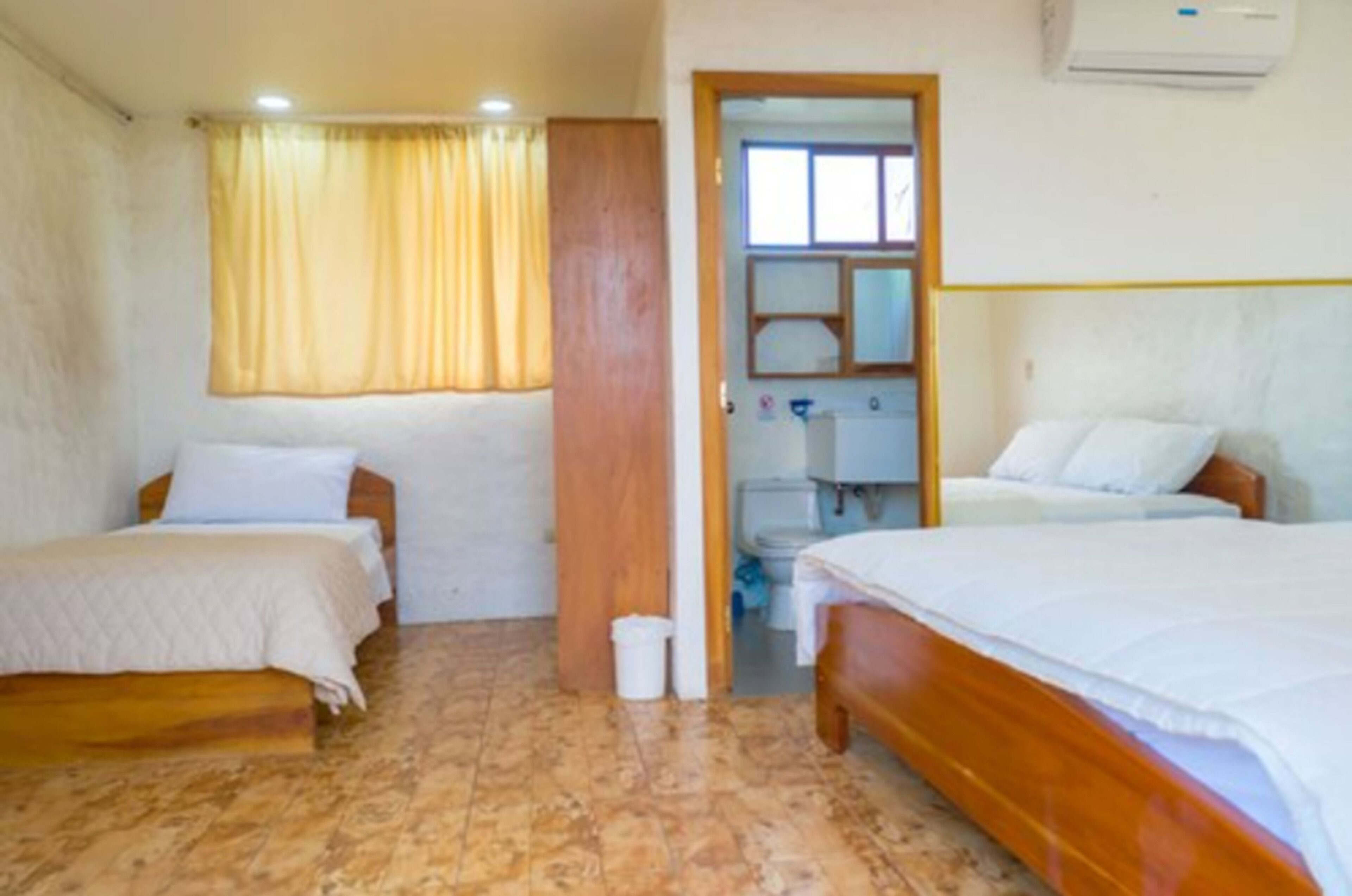 A room with two beds, a wooden wardrobe, a bathroom with a toilet and sink, and a window with curtains.