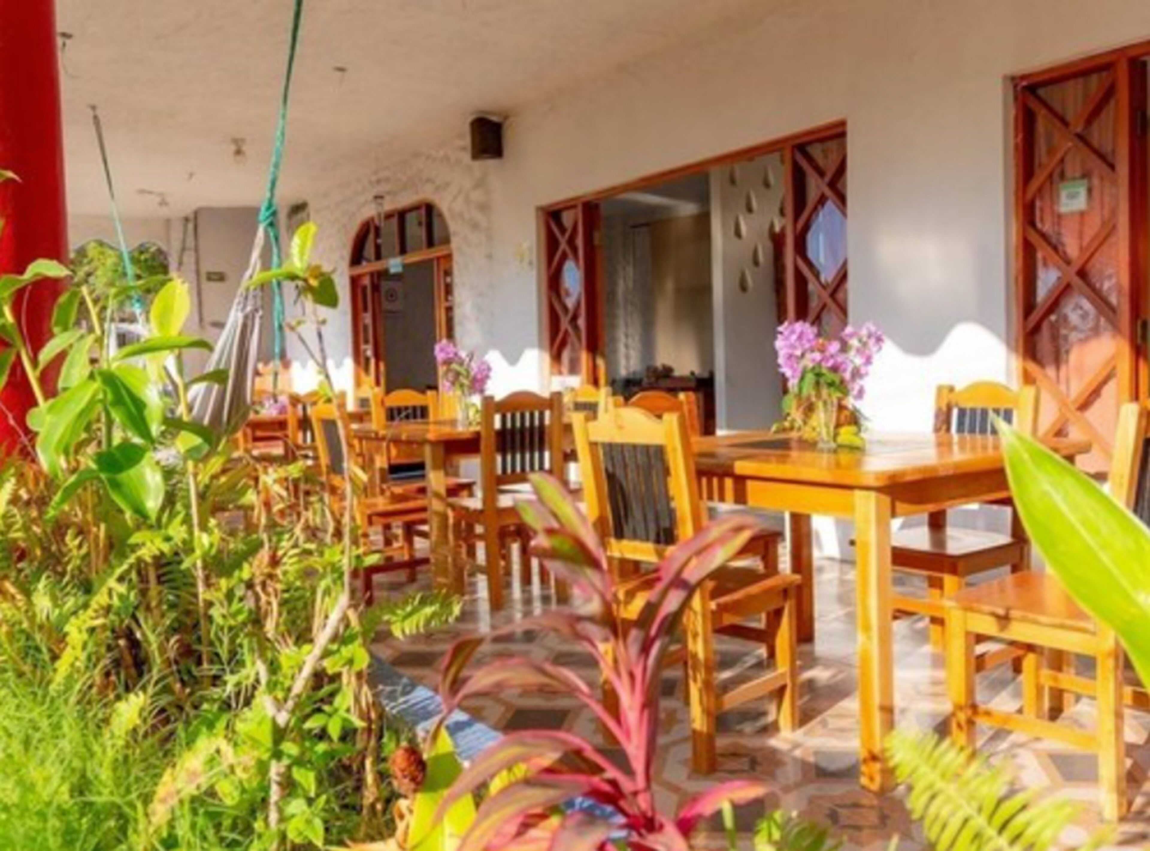 A covered outdoor dining area with wooden tables and chairs, surrounded by tropical plants and a tiled floor.