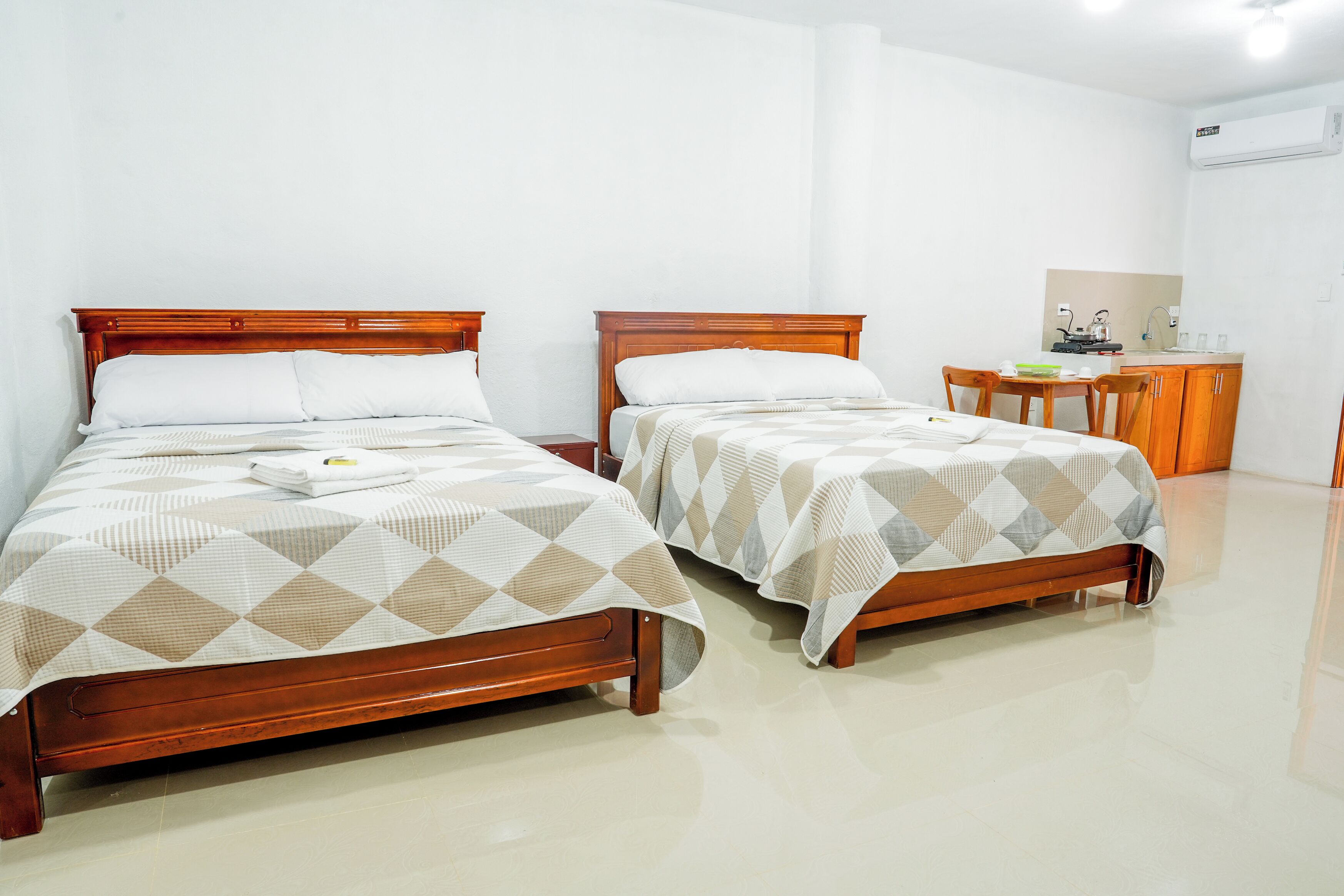 A hotel room with two wooden beds, a small desk with a chair, and a kitchenette area.