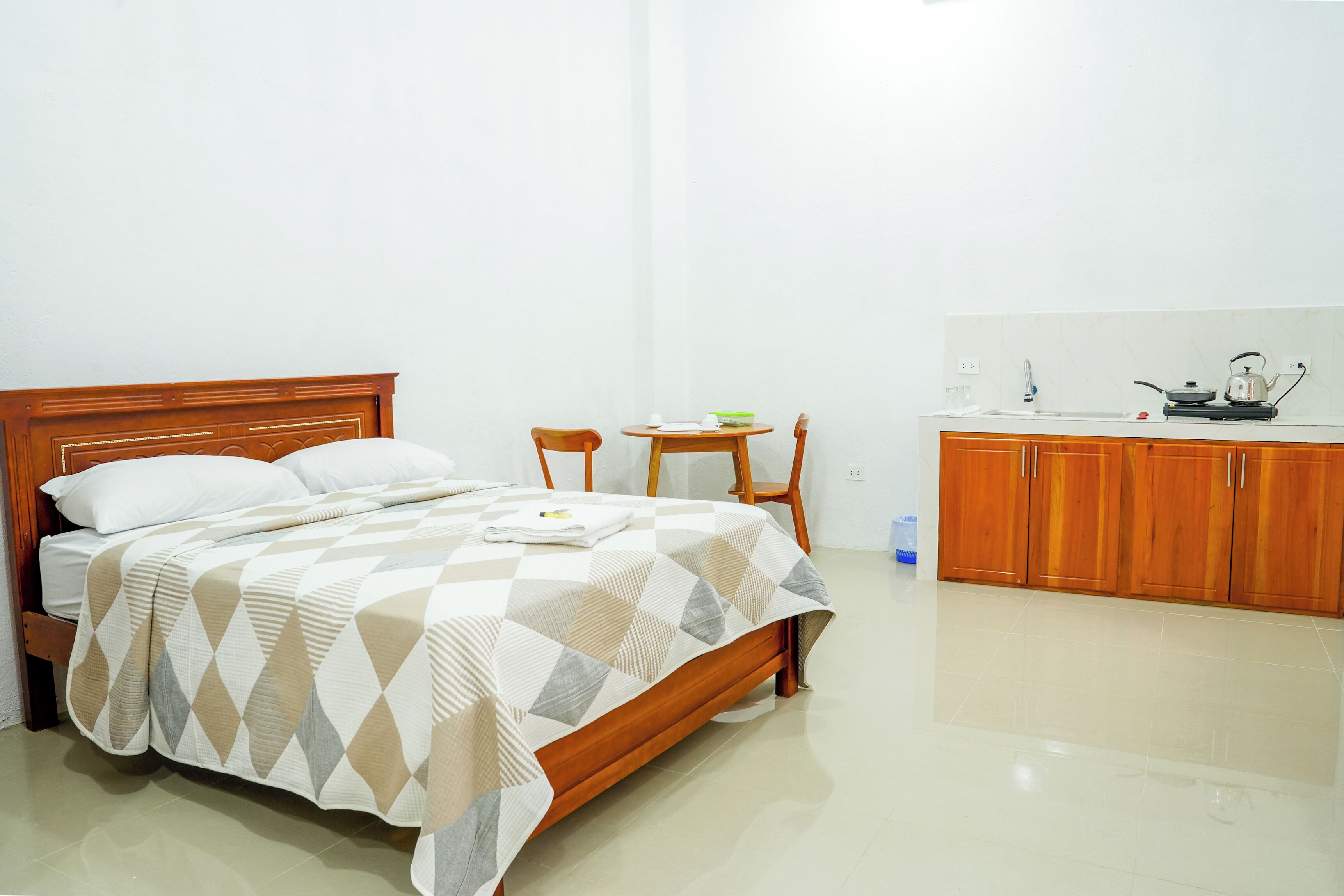 A bedroom with a wooden bed, a small round table with chairs, and a kitchenette area with cabinets and a kettle.