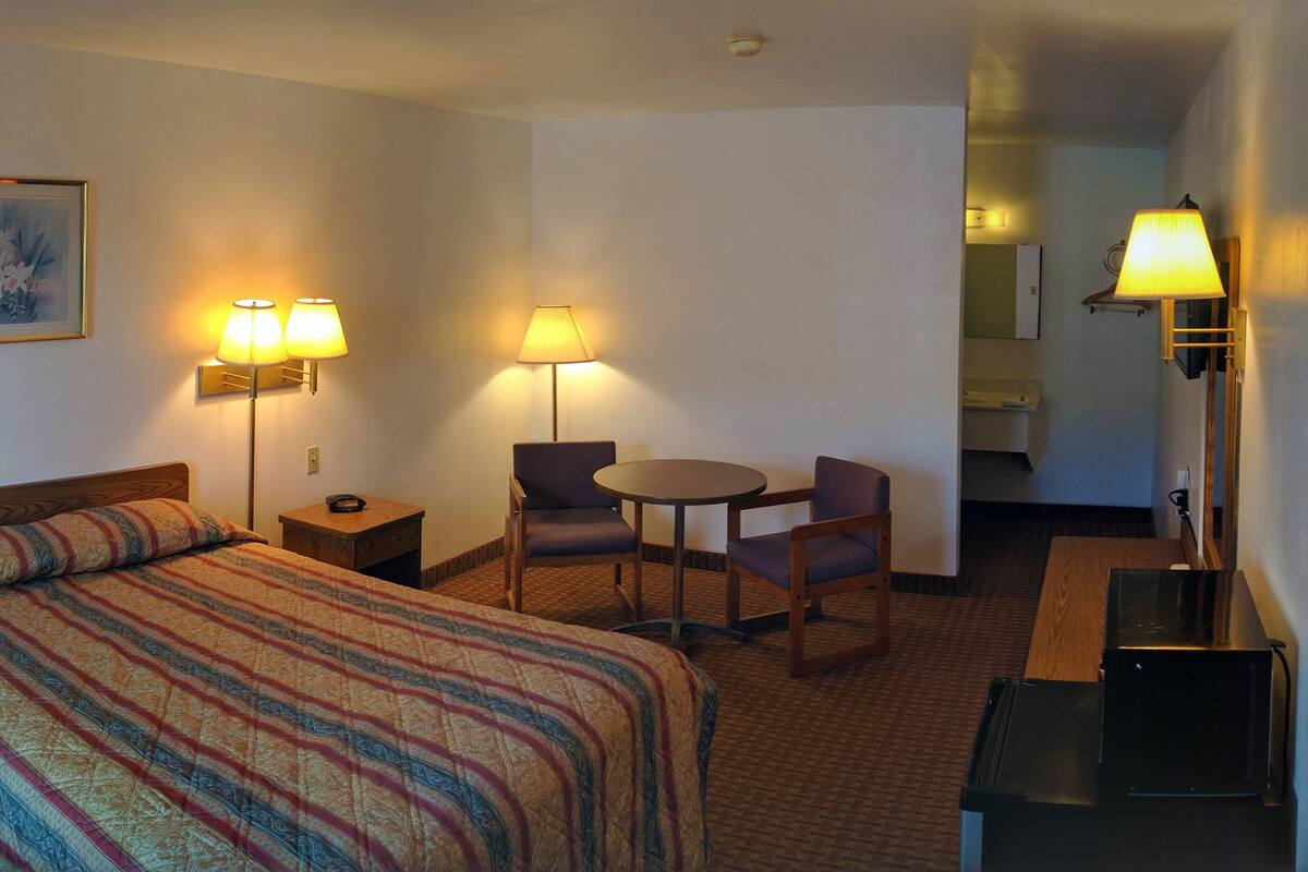 A hotel room with a bed, two bedside lamps, a small table with a chair, a painting on the wall, and a mirror.