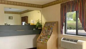 A reception area with a counter, a poster board, a window with curtains, and a wall-mounted air conditioner. - Value Inn Harrisburg - York (Etters)