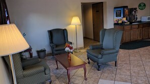 A hotel lobby with two patterned armchairs, a wooden coffee table with a vase of flowers, a floor lamp, and a breakfast bar with a coffee machine. - Value Inn Harrisburg - York (Etters)