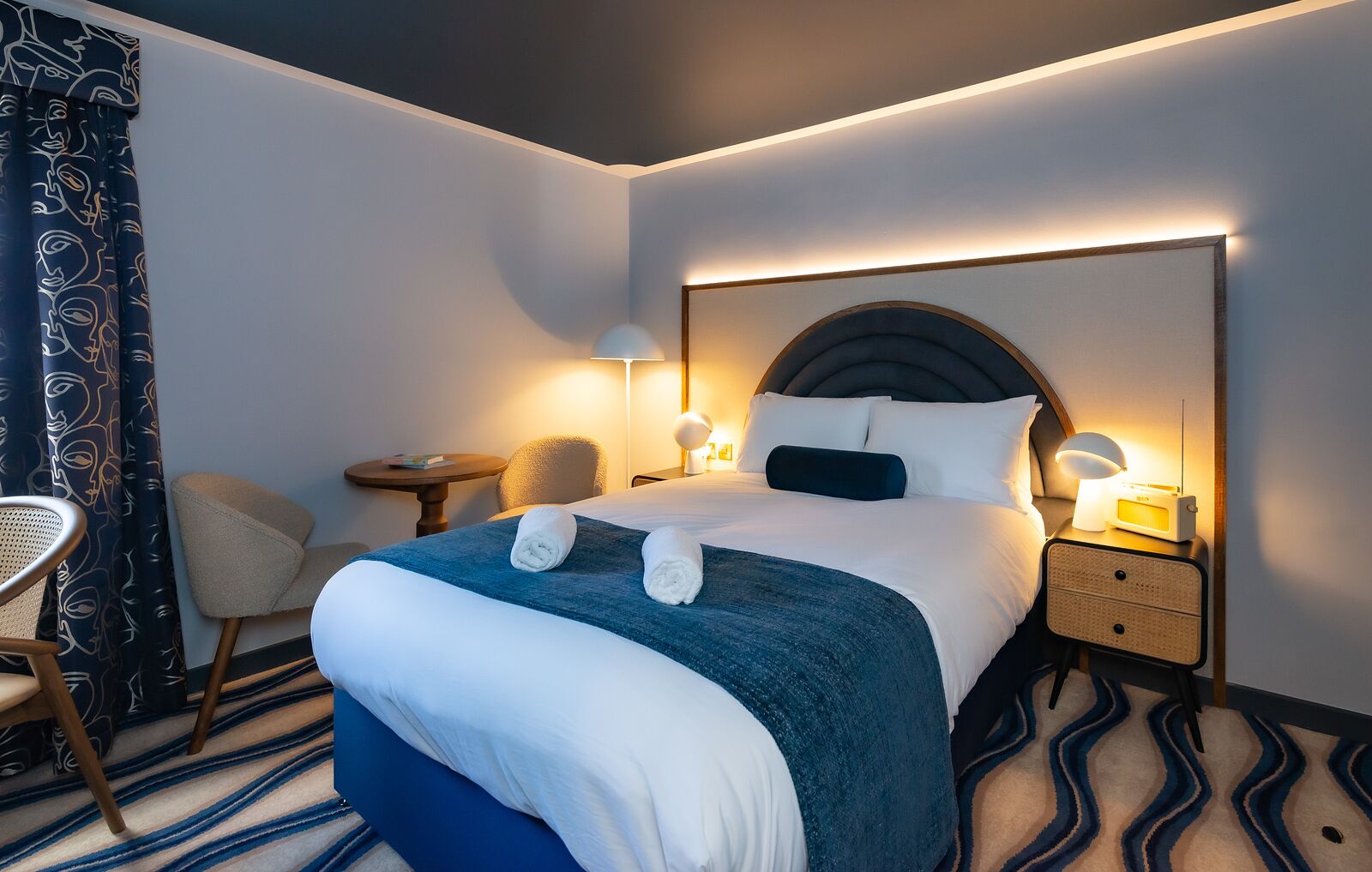 A hotel room with a large bed, two bedside tables, a chair, a lamp, and a patterned curtain.