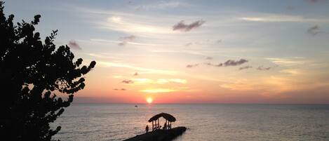Un coucher de soleil sur une mer calme, avec une jetée qui s’avance dans l’eau, une structure à toit de chaume sur la jetée et un arbre sur la gauche.