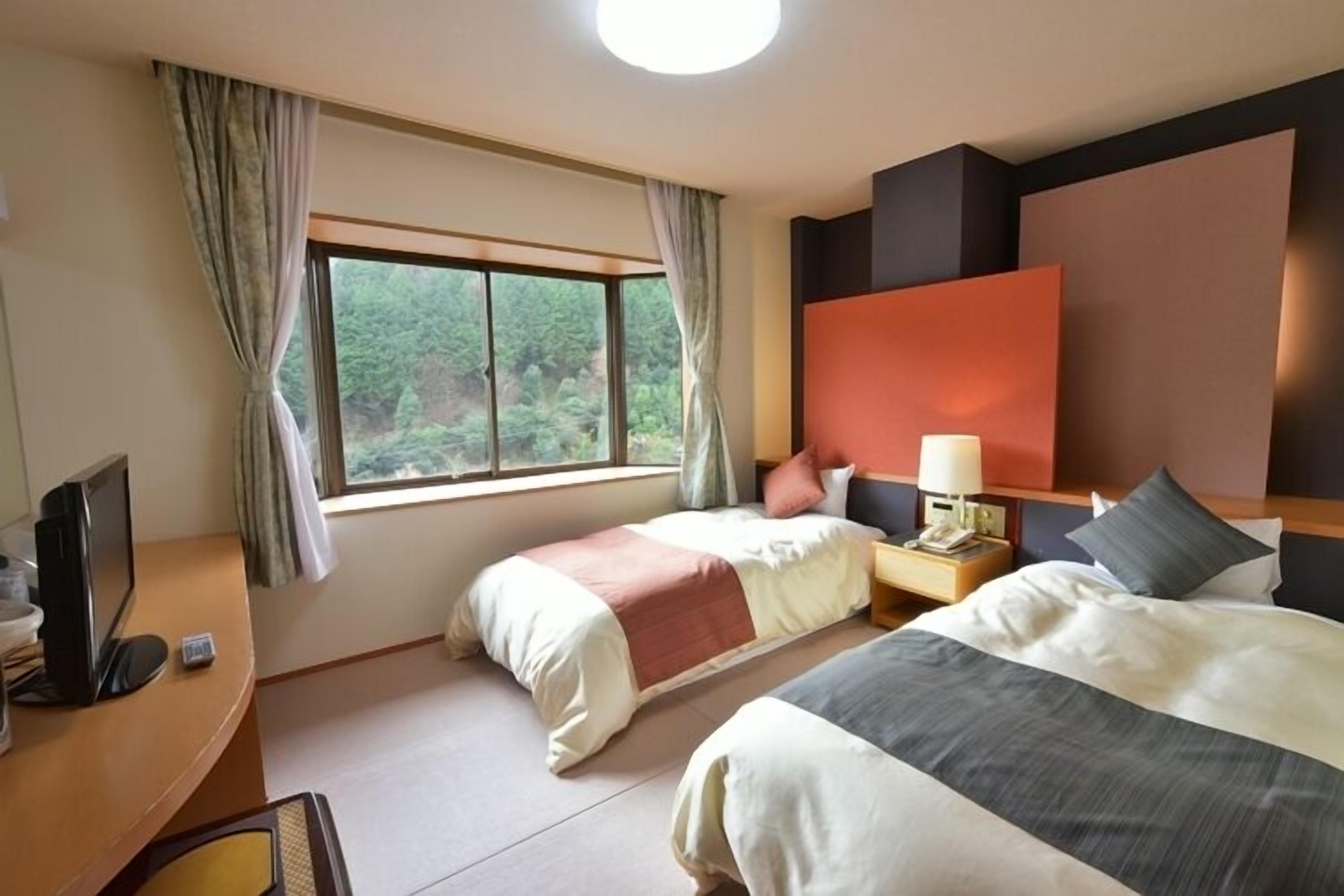 A hotel room with two beds, a desk with a computer, a nightstand with a lamp, and a view of greenery outside.