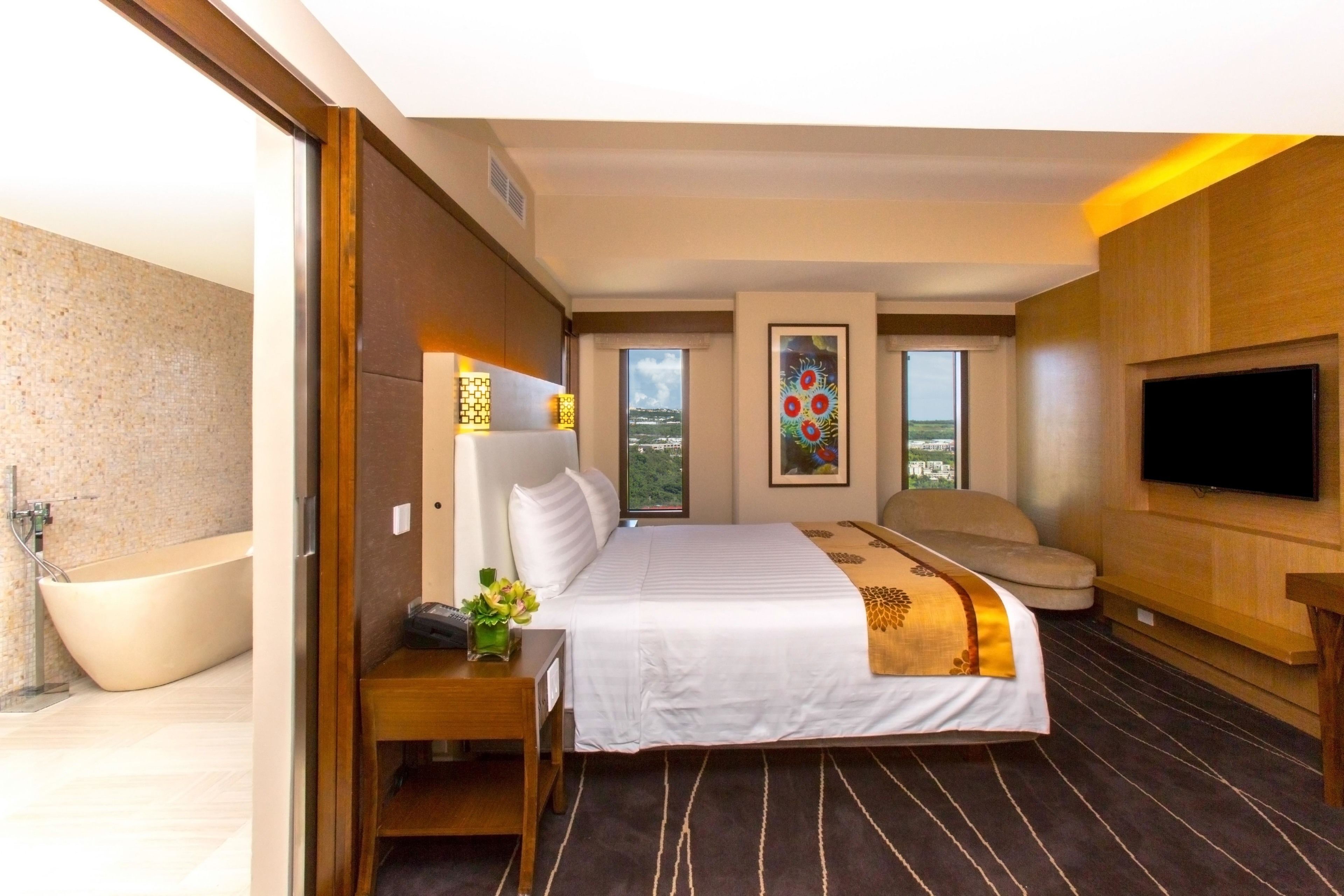 A hotel room with a large bed, a flat-screen TV, a bathtub, and a view of the city.