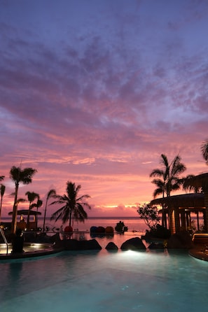 A resort pool area with palm trees, a thatched-roof structure, and a view of the ocean at sunset.