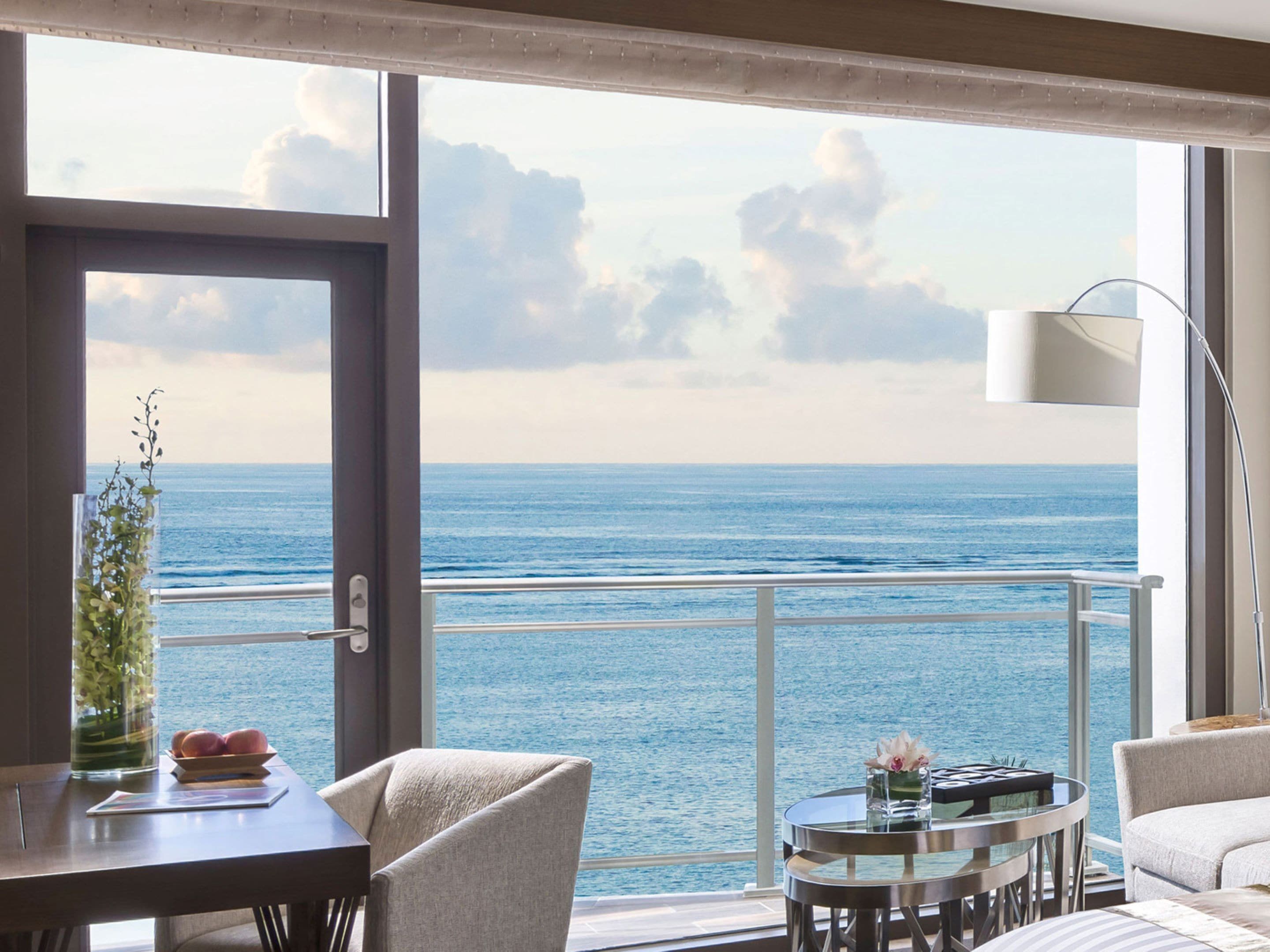 A room with a view of the ocean, a dining table with a vase of flowers, and a round table with a glass top.