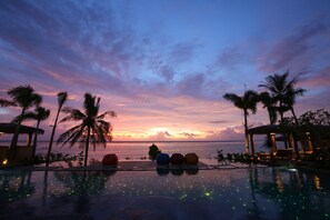 A poolside view with palm trees and a sunset over the ocean.