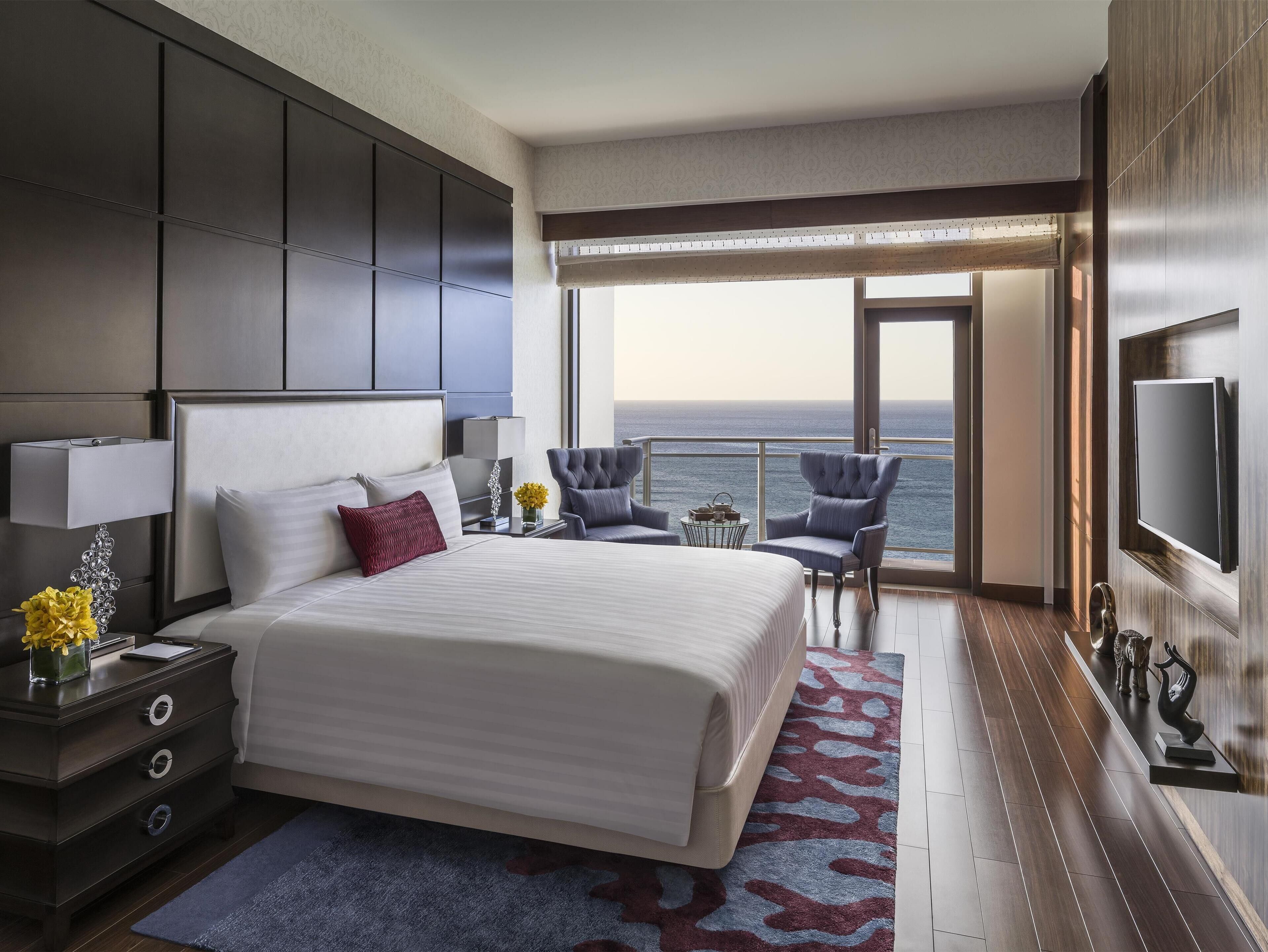 A modern hotel room with a large bed, a desk with a lamp, a chair, a small table with flowers, and a view of the ocean through a balcony.