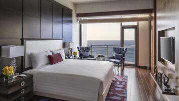A modern hotel room with a large bed, a desk with a lamp, a chair, a small table with flowers, and a view of the ocean through a balcony.