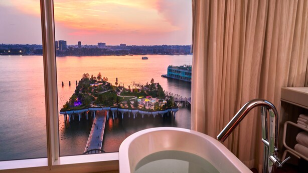 Une chambre d’hôtel avec vue sur un parc au bord de l’eau, particulièrement belle au coucher du soleil, équipée d’une baignoire et d’un rideau.