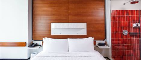 Quarto de hotel com cabeceira de madeira, cama branca com duas almofadas, mesa de cabeceira com telefone e área de chuveiro com azulejos vermelhos.