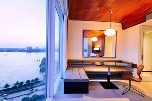 A modern hotel room with a large window overlooking a body of water, a built-in bench, a round table, and a unique ceiling design.