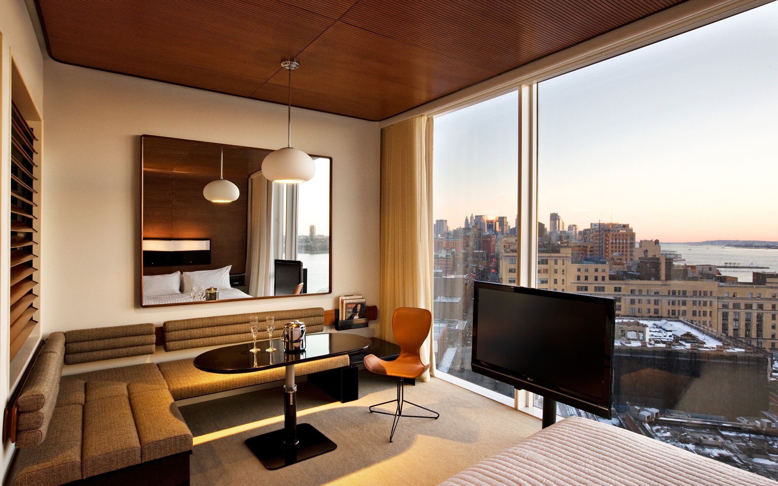 A modern hotel room with a large window, a sofa, a dining table, and a TV.