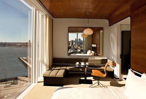 A modern hotel room with a large window offering a city view, a cozy seating area, and a dining space.