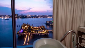 Une salle de bain avec une baignoire, une fenêtre donnant sur un paysage urbain au crépuscule et un rideau.