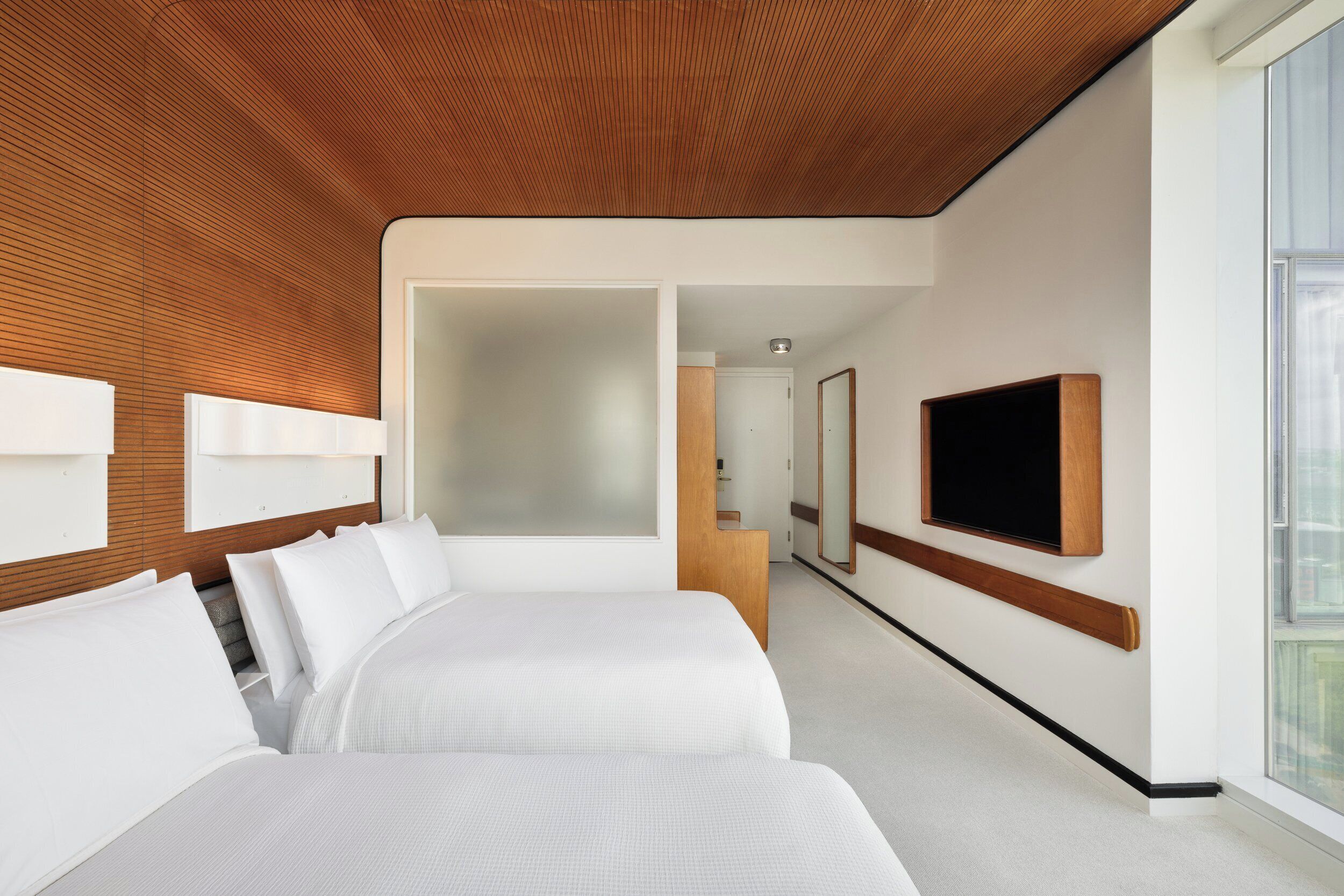 A modern hotel room with two beds, a wooden ceiling, a TV mounted on the wall, and a large window.