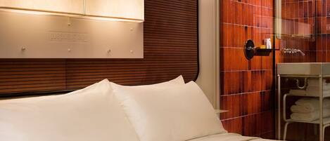A hotel room with a bed, a wooden headboard, a wall-mounted light, a glass shower enclosure, and a towel rack.