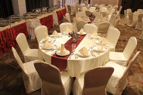 Un salón de banquetes con mesas redondas dispuestas para un evento formal, cada una con manteles blancos, cubertería y cristalería.