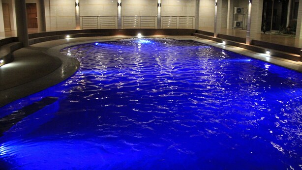 Ein großzügiges Hallenbad mit einem blau beleuchteten Wasserspiel, umgeben von moderner Architektur mit mehreren Sitzbereichen und einem überdachten Gang.