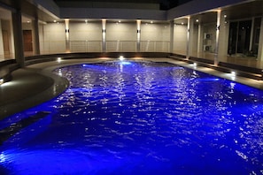 Una amplia piscina interior con una fuente iluminada en azul, rodeada por un diseño arquitectónico moderno con varias zonas de estar y un paseo cubierto.