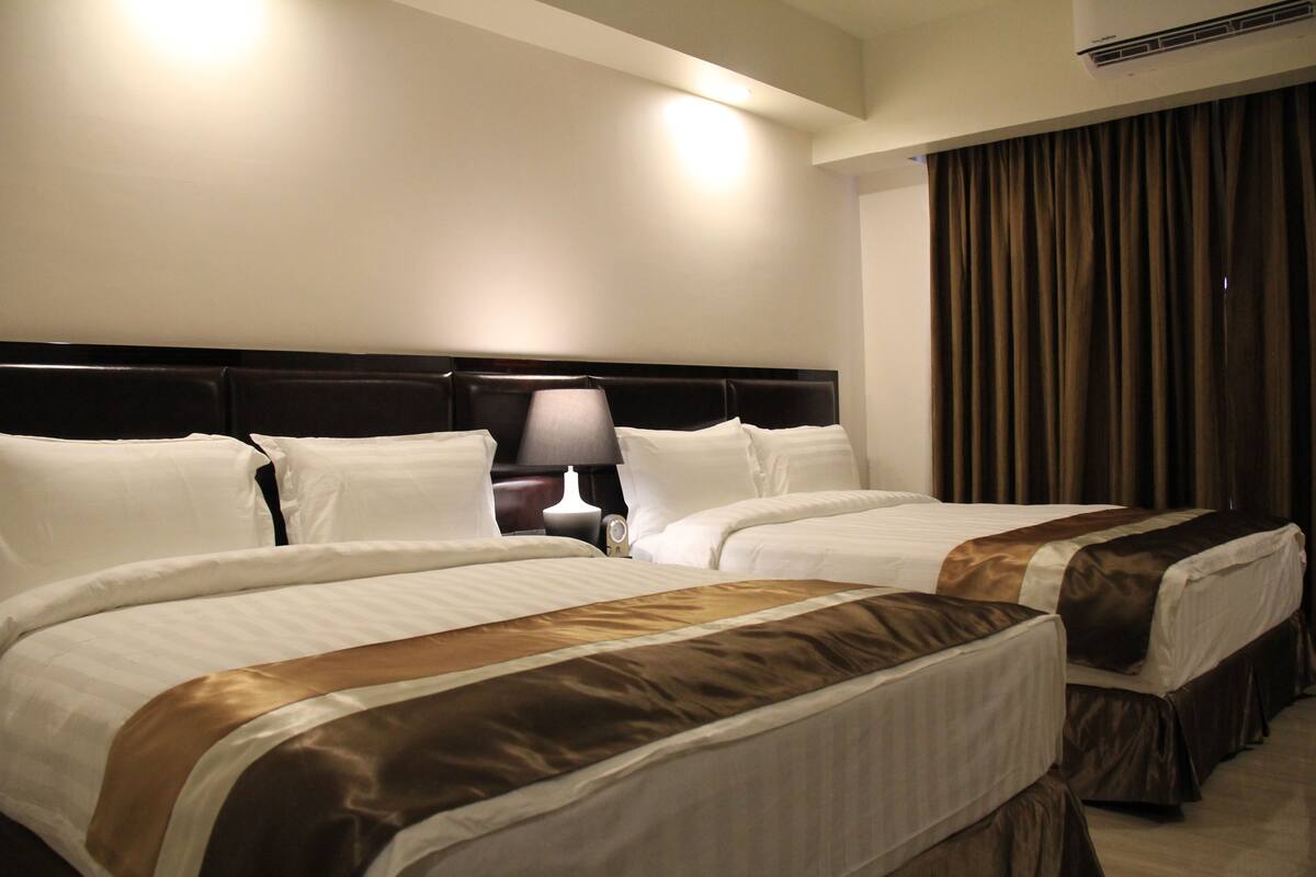 Une chambre d’hôtel avec deux lits, une table de chevet avec une lampe et une grande fenêtre avec des rideaux.