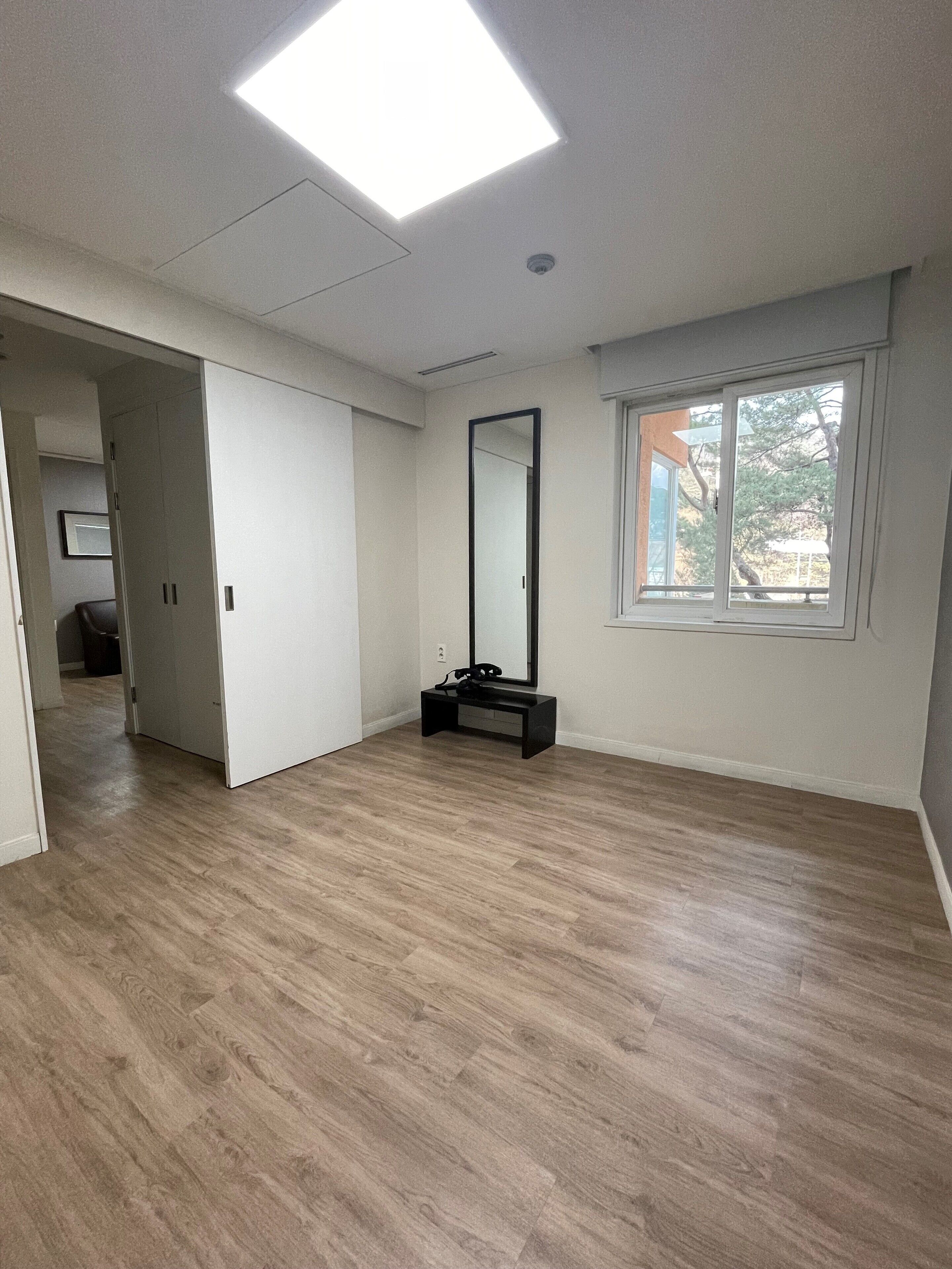 A spacious room with a large window, a mirror, and a wooden floor.