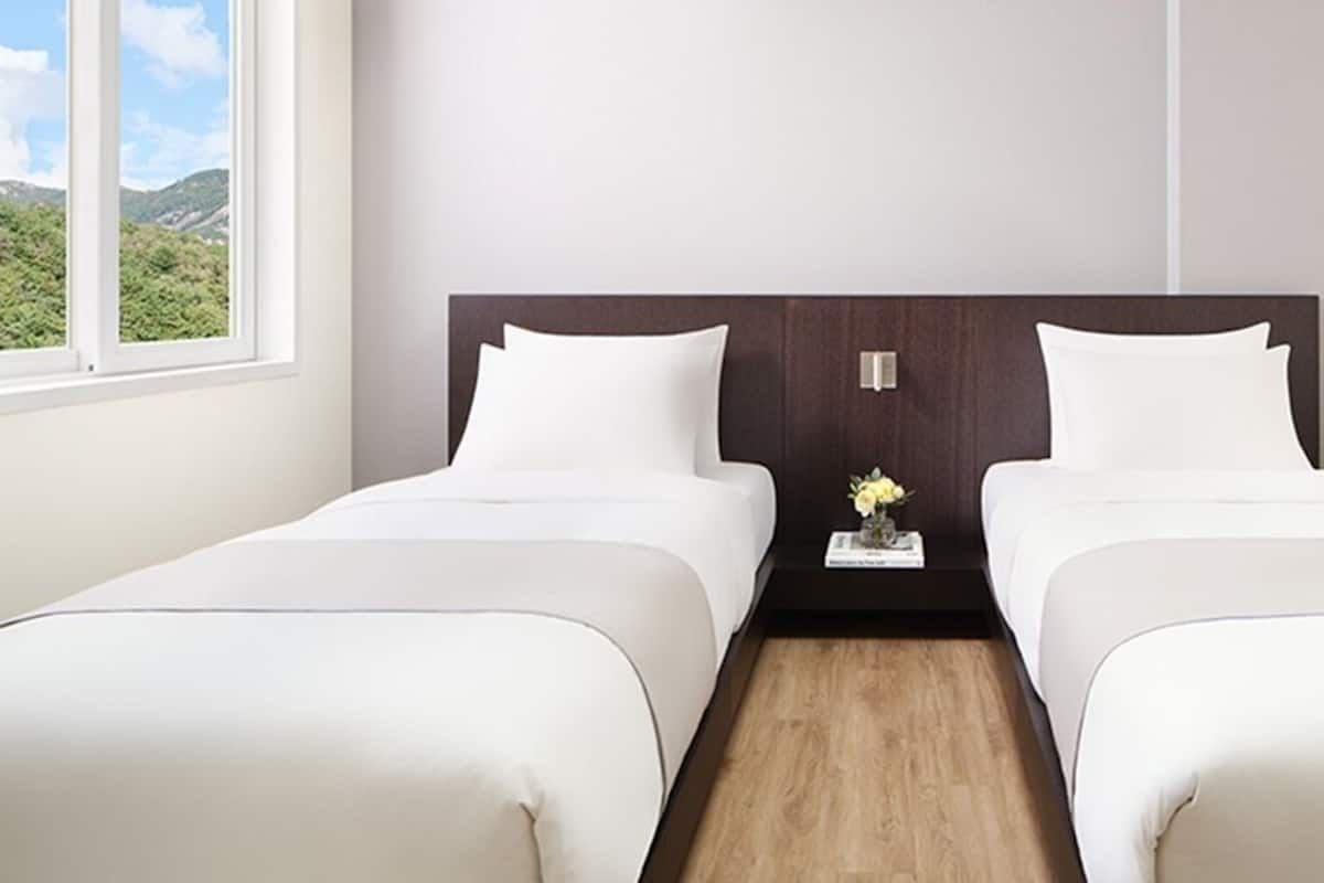 A hotel room with two beds, a nightstand with a vase of flowers, and a window with a view of greenery.
