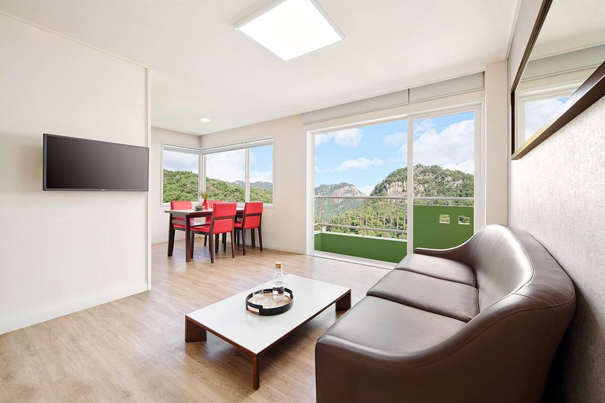A modern living room with a flat-screen TV, a brown leather sofa, a dining area with red chairs, and large windows offering a view of greenery.