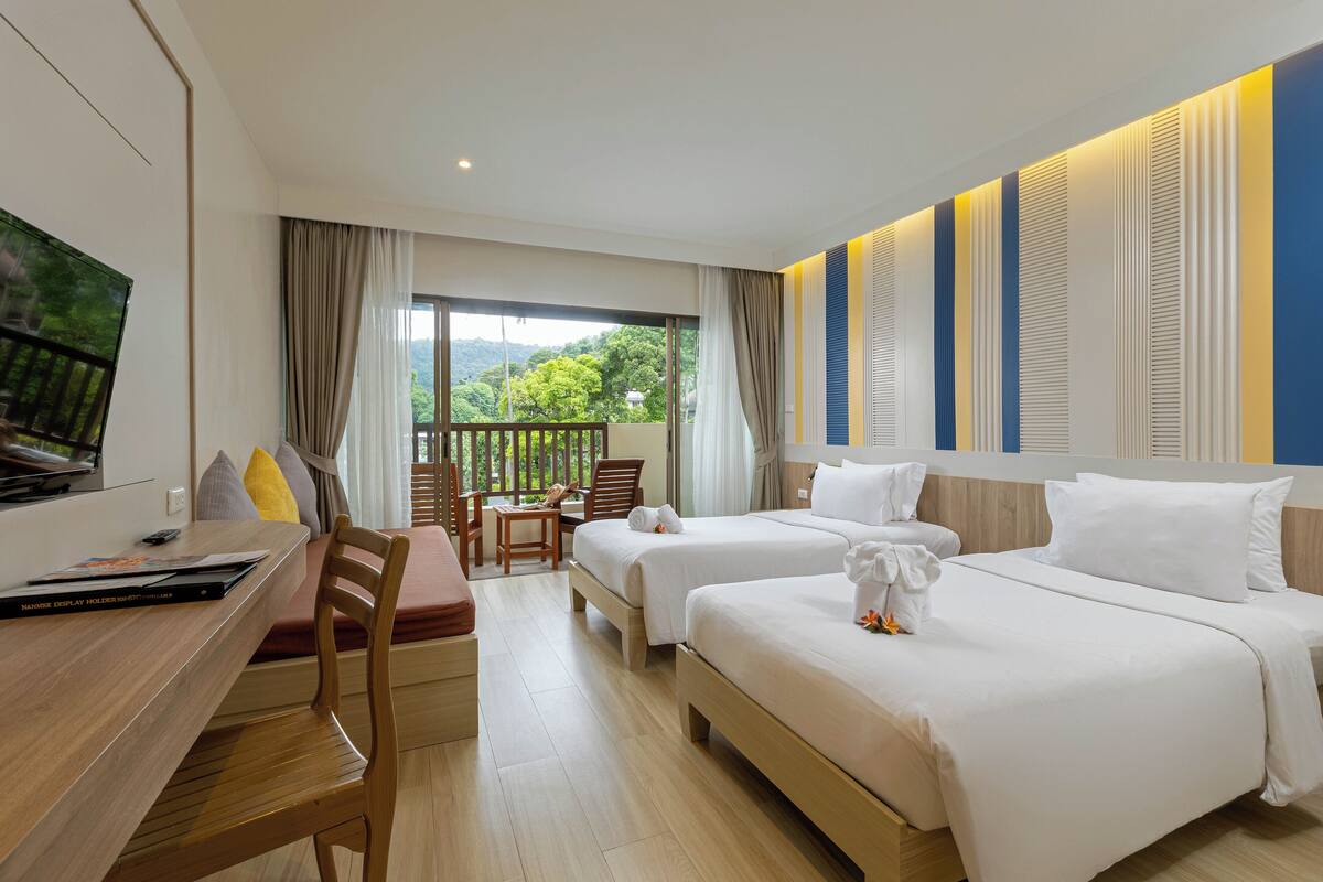 A hotel room with two beds, a desk with a chair, a TV, and a balcony with a view of greenery.