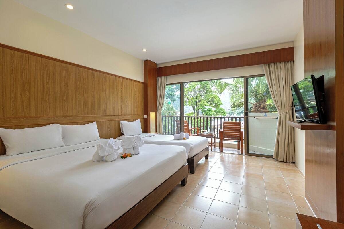 A hotel room with two beds, a TV, and a balcony with a view of greenery.