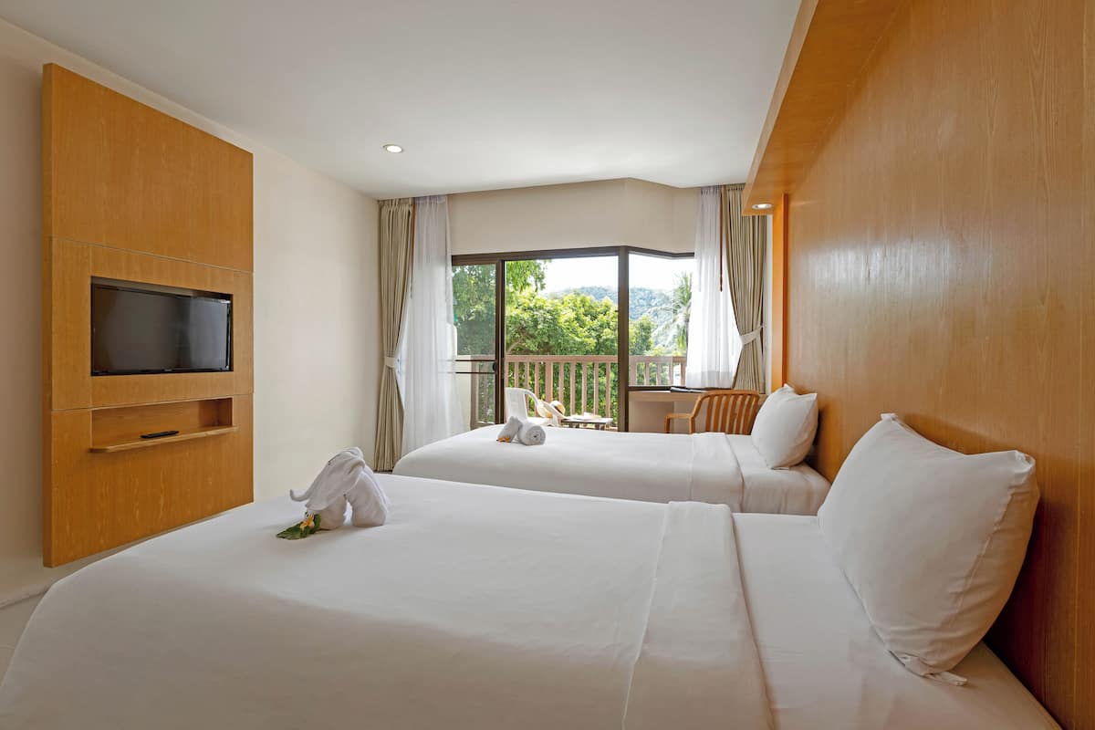 A hotel room with two beds, a TV mounted on the wall, and a balcony with a view of greenery.