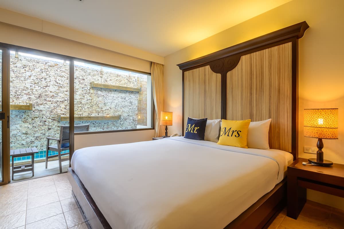 A large bed with two pillows labeled "Mr." and "Mrs.", a nightstand with a lamp, a window with curtains, and a view of a stone wall and outdoor seating area.