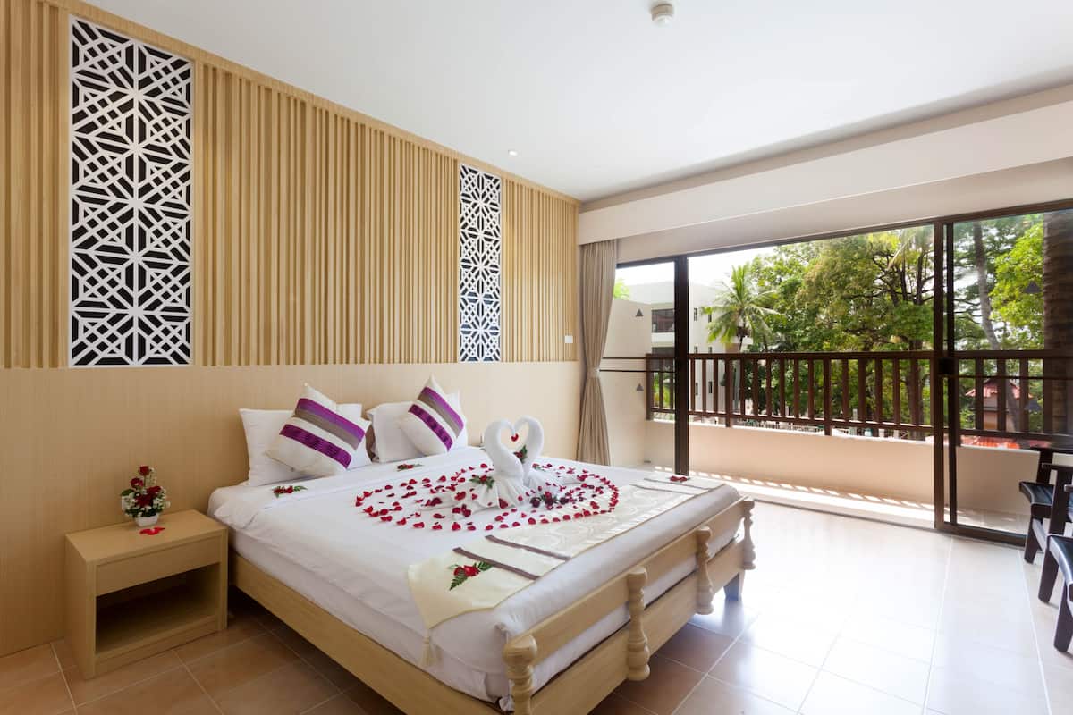 A hotel room with a bed, a nightstand, a balcony with a view of trees and a building.