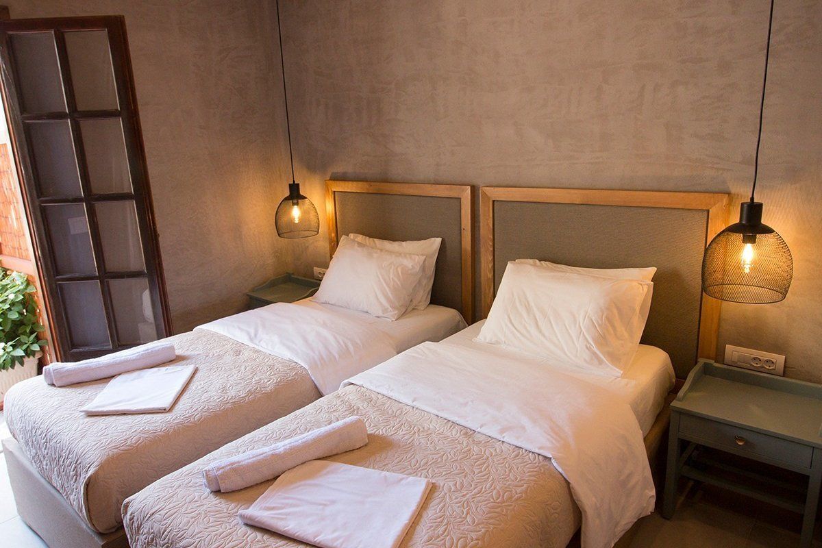 Habitación de hotel con dos camas, cada una con cabecero de madera, ropa de cama blanca y una mesita de noche con lámpara.