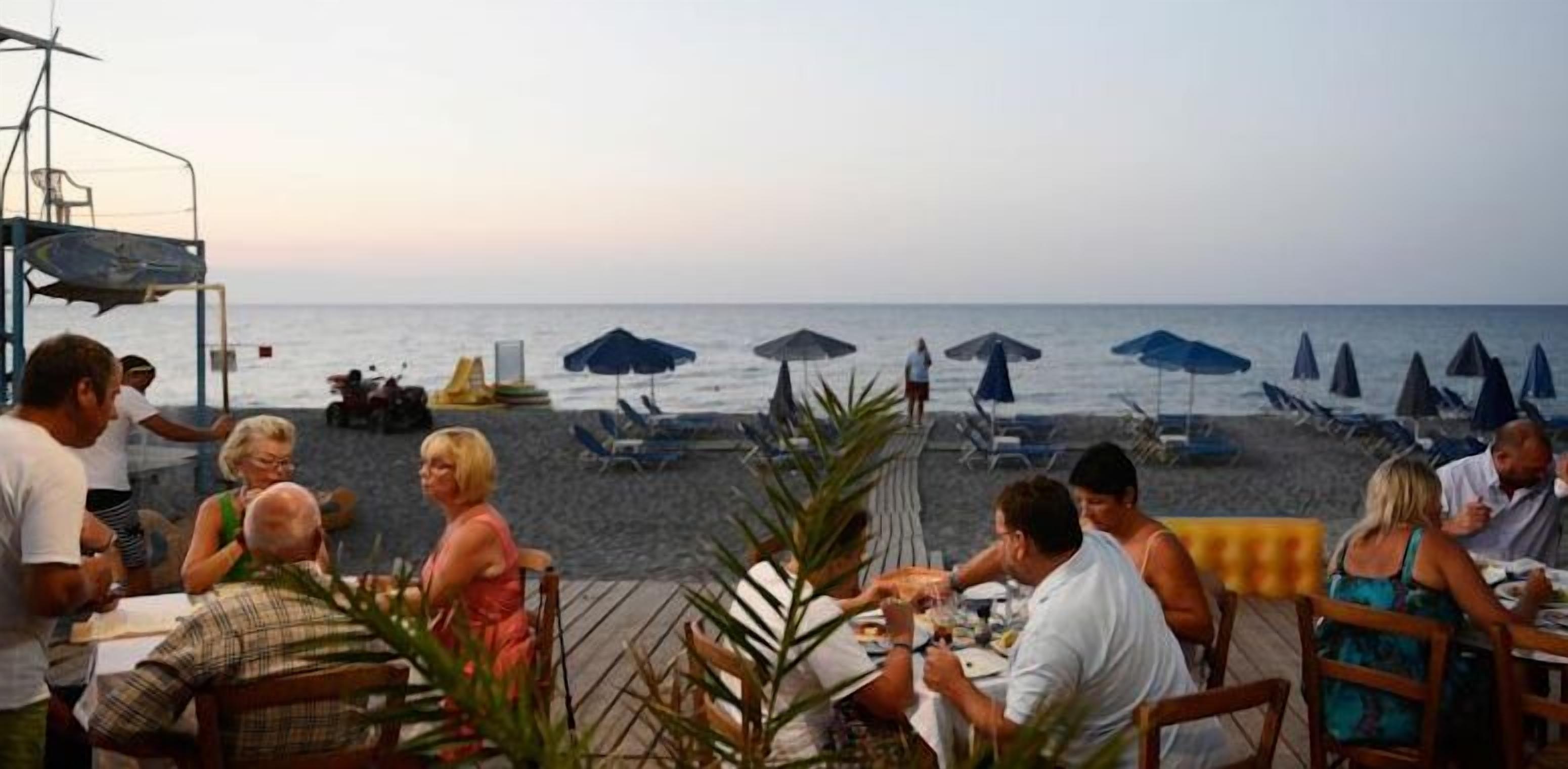 A beachside restaurant with outdoor seating overlooking the sea, umbrellas, and lounge chairs.