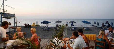 A beachside restaurant with outdoor seating overlooking the sea, umbrellas, and lounge chairs.