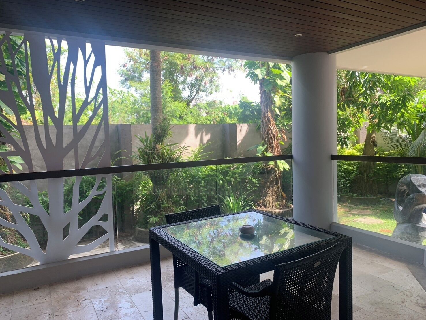 A covered patio with a glass table and chairs, surrounded by a white decorative fence and lush greenery.