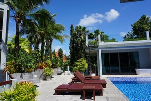 Un área de piscina moderna al aire libre, con sillones de descanso y una variedad de plantas tropicales.