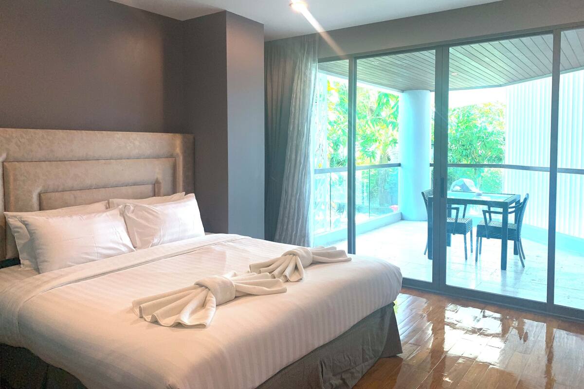 Une chambre d’hôtel avec un grand lit, un balcon doté d’une table et de chaises, et une vue sur la verdure environnante.