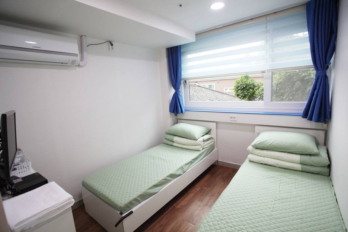 Una habitación pequeña y limpia con dos camas individuales, un escritorio con computadora y una ventana con cortinas.