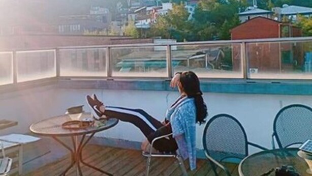 Una persona sentada en una silla de terraza con vista a una piscina, y en la distancia, una colina y una torre.