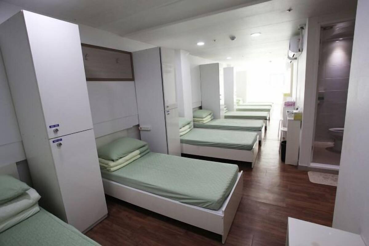 Un pasillo con varias camas individuales, cada una con colchón verde y estructura blanca, que conduce a un área de baño.
