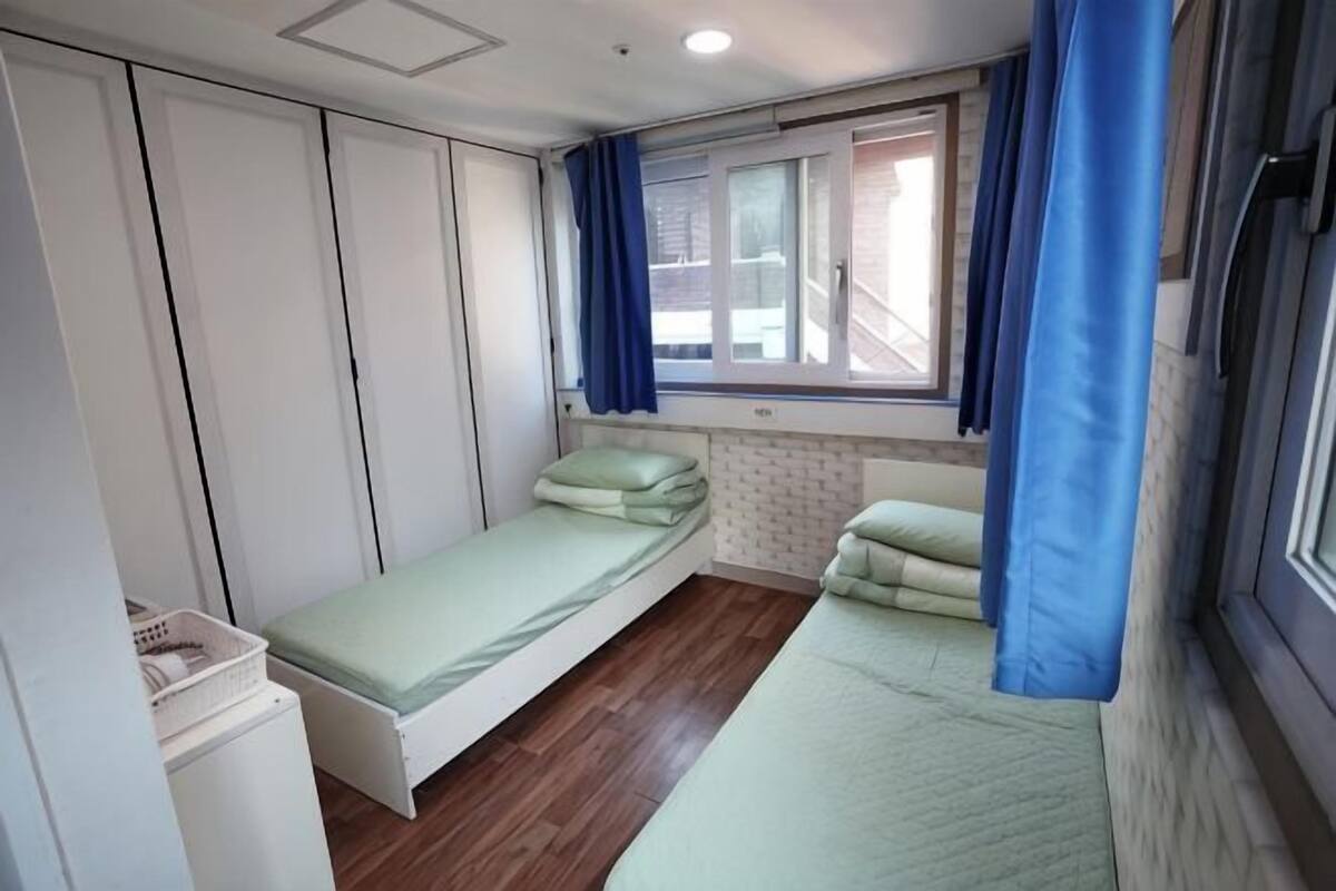 Una habitación pequeña y funcional con dos camas individuales, un armario y una ventana con cortinas.