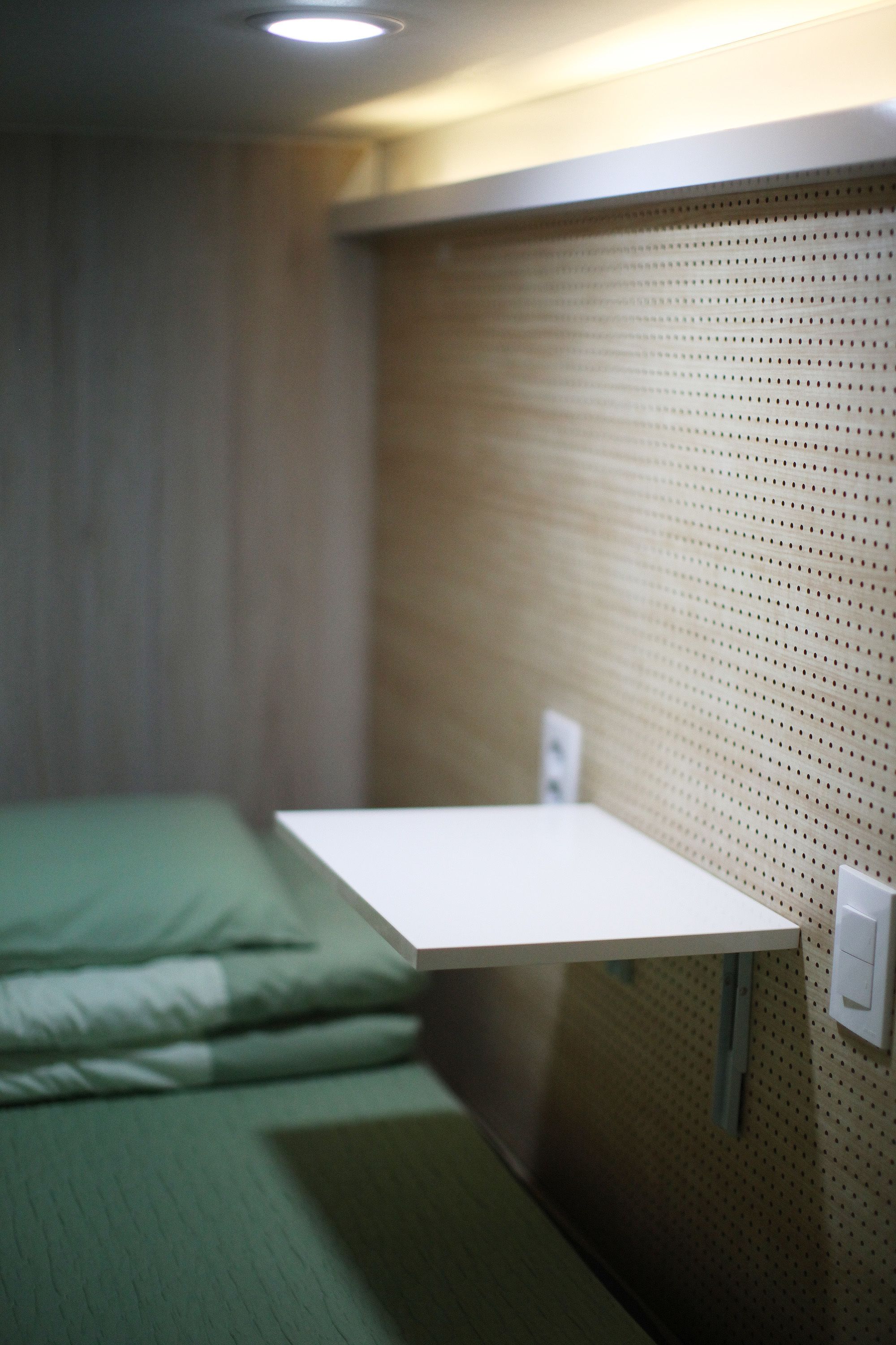 A small, single-bed room with a wooden wall and a white shelf.