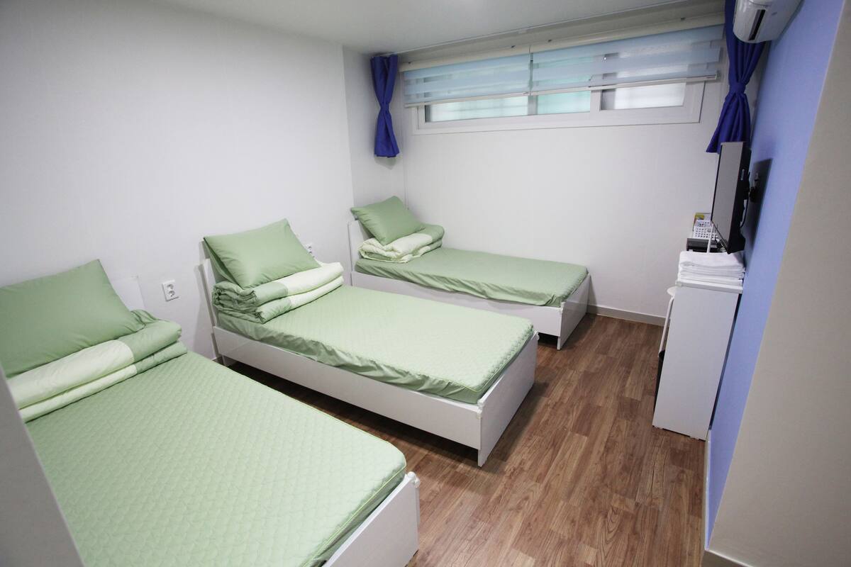 Una habitación con tres camas individuales, cada una con ropa de cama verde, un pequeño armario blanco y una ventana con cortinas azules.